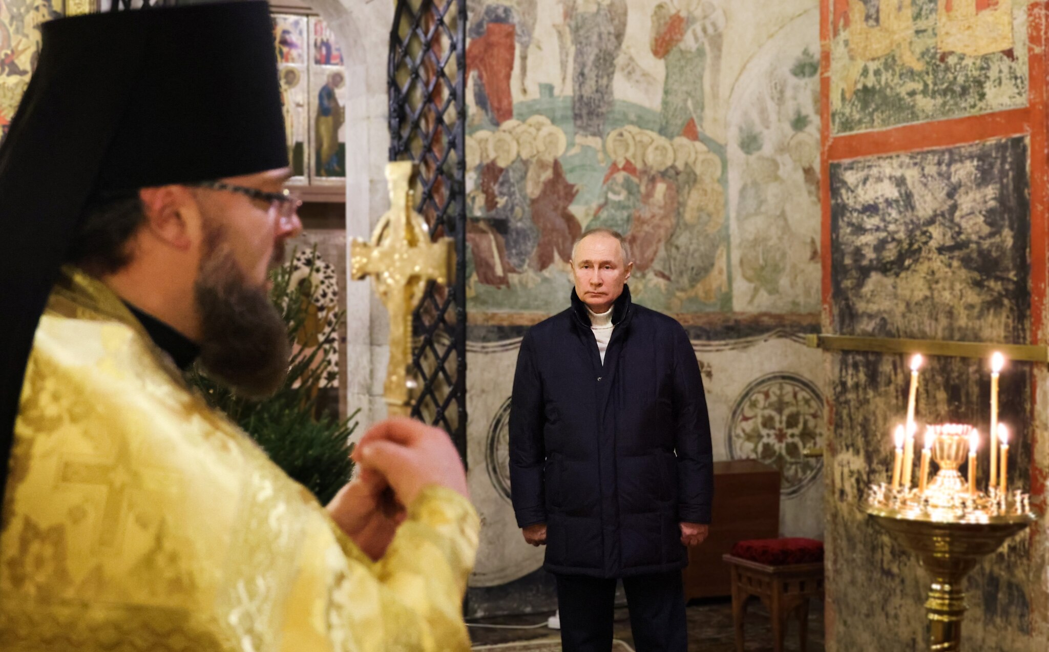 Vladimir Putin stands stiffly in a corner, staring straight ahead as a priest gives a blessing holding a cross.