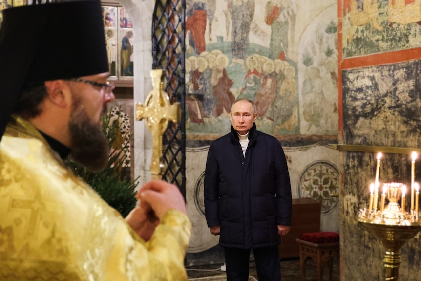 Vladimir Putin stands stiffly in a corner, staring straight ahead as a priest gives a blessing holding a cross.