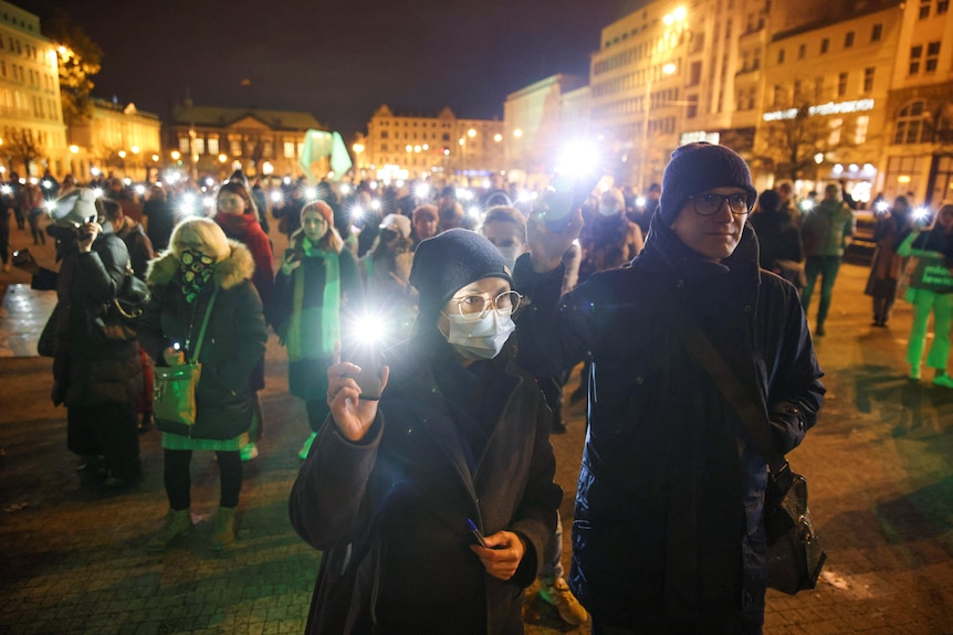 People hold candles and torches as they protest in an open public space surrounded by illuminated buildings.