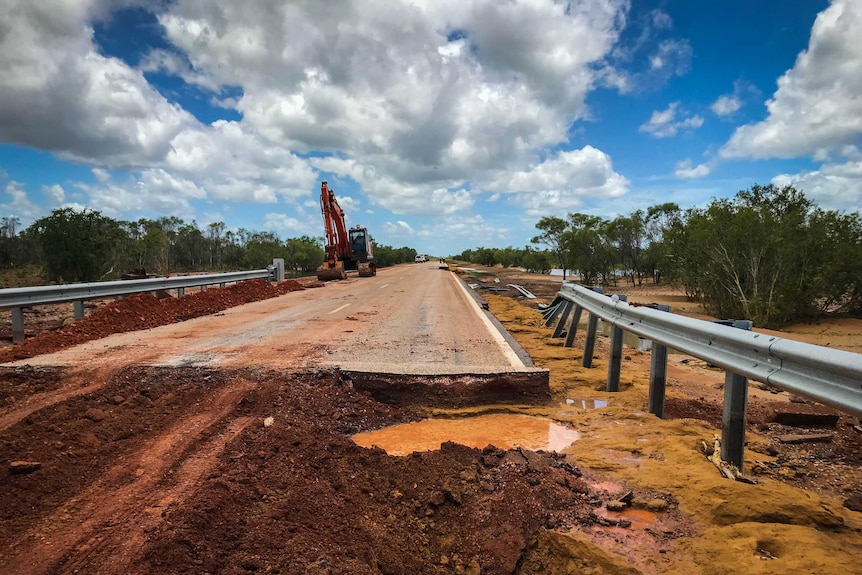 A bitumen road destroyed by flooding, with red dirt in the foreground, and a digger further down the road.