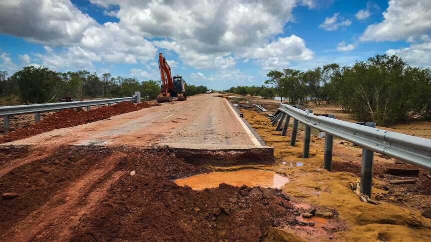 A bitumen road destroyed by flooding, with red dirt in the foreground, and a digger further down the road.