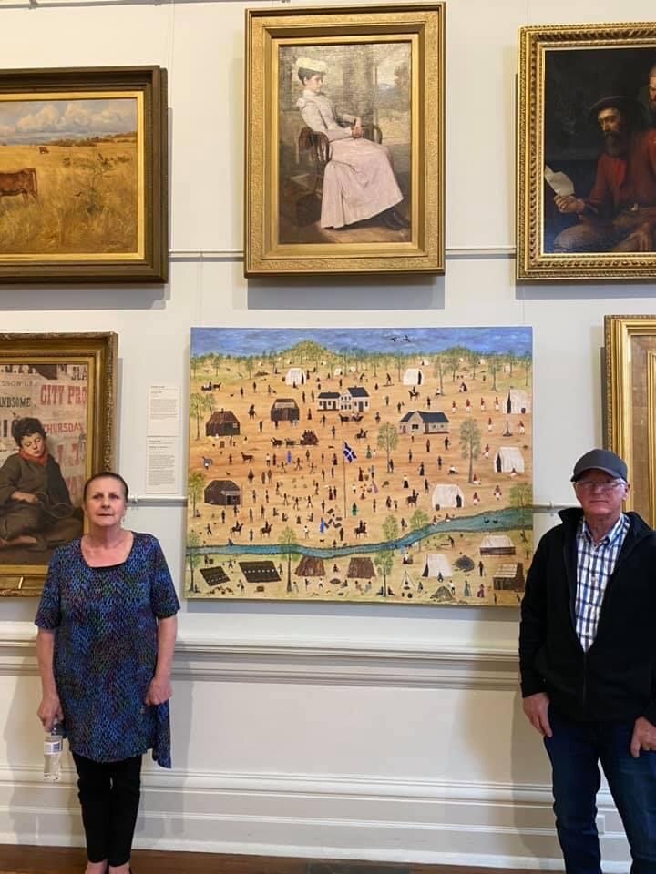 A woman and a man stand in front of painting in an art gallery.