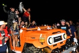 An orange car is full of happy people, one is holding a fish in the air. The car is on the beach and it is dark. 