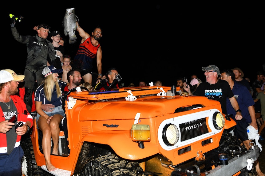 An orange car is full of happy people, one is holding a fish in the air. The car is on the beach and it is dark. 