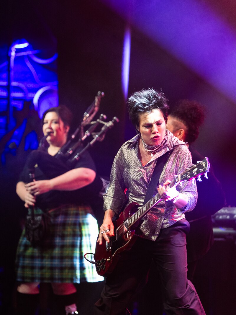 Sarah McLeod plays electric guitar on stage in the foreground with Camille Layt on bagpipes in the background. 