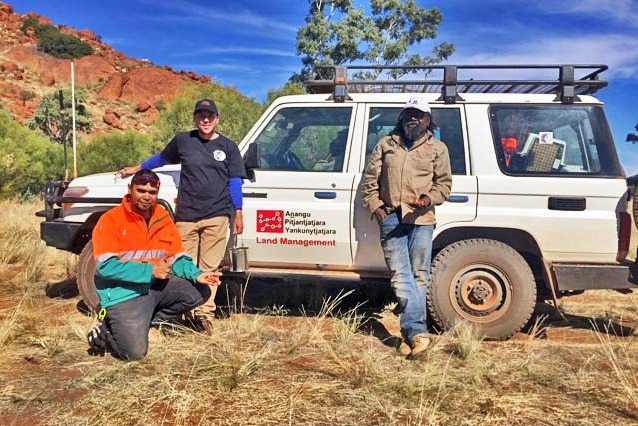 Three men standing near a white four-wheeled drive among dry grass, with a rocky hill and blue sky in the background.