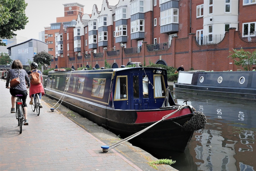 A picture of a dark blue canal boat in the water as two cyclists ride on the footpath