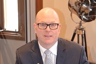 John Crowley, wearing glasses and a suit, looks at the camera while sitting behind a desk.