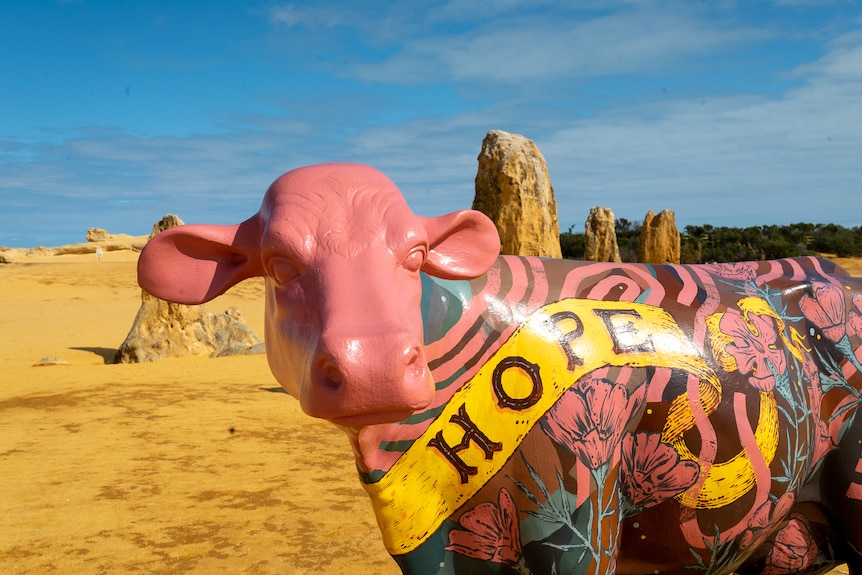 A pink cow sculpture stands in the desert. 