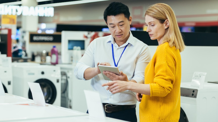 A woman wearing a yellow sweater negotiates at a whitegoods store with a salesman of Asian appearance. 