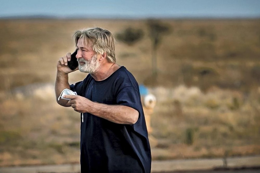 a man with grey-brown hair and beard makes a phone call in desolate dry countryside 