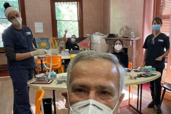 A man with grey hair and wearing a medical mask takes a selfie with four others wearing masks and scrubs in background