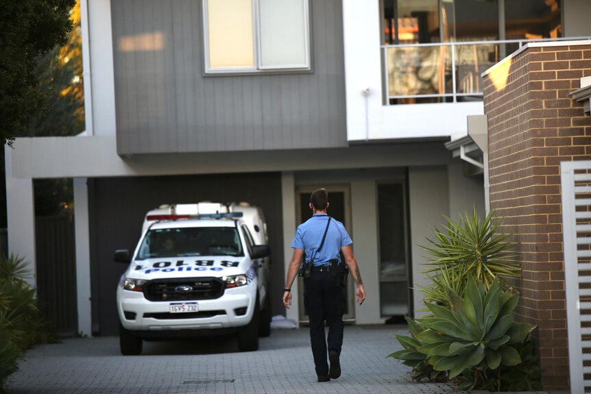 A police officer walks towards a unit complex down a driveway with a police vehicle parked against the building.