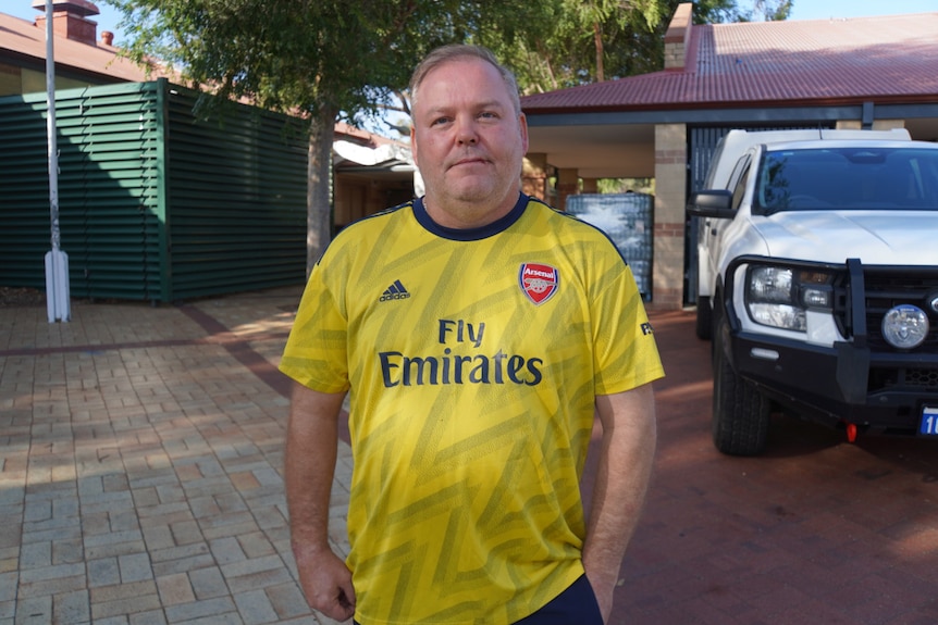 A mid-shot of a middle-aged man wearing a yellow Arsenal shirt, standing in front of a 4WD and a building.