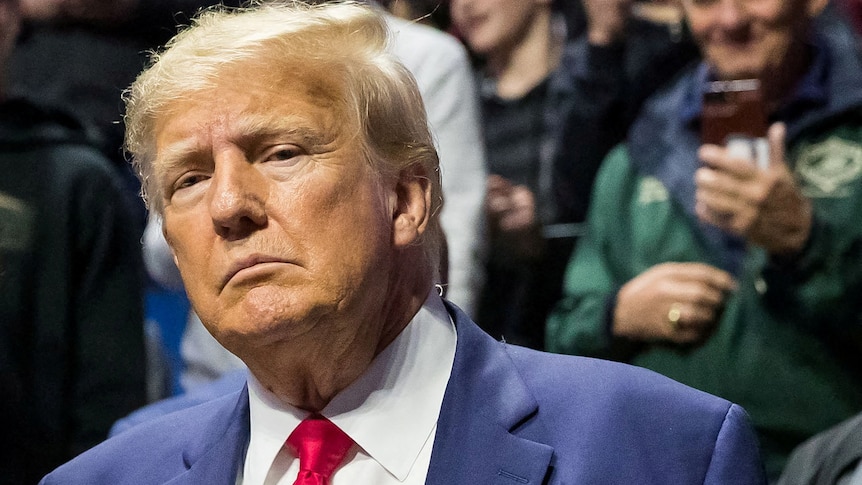 Donald Trump dressed in a blue suit and red tie sits at a game.