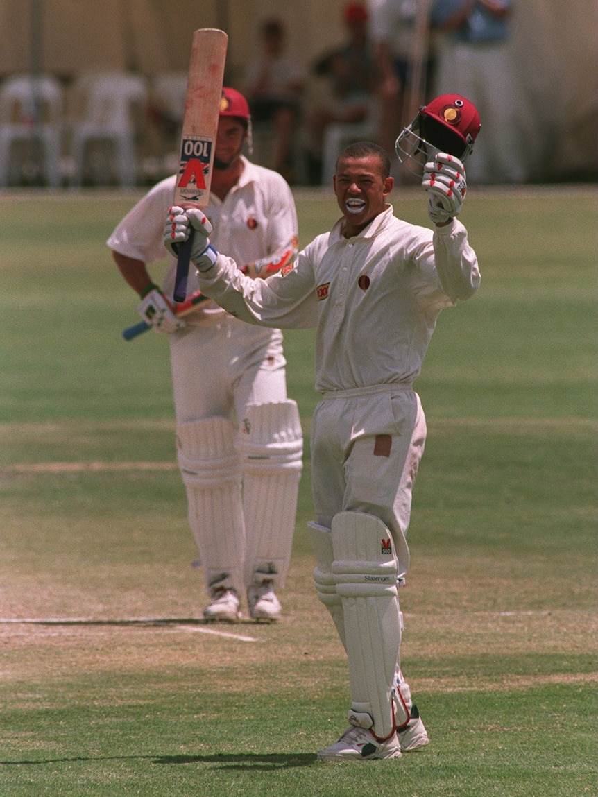 A man in cricket whites raises his bat and helmet in celebration.