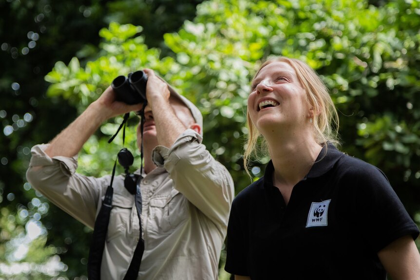 Woman standing and looking up at trees smiling next to man with binoculars