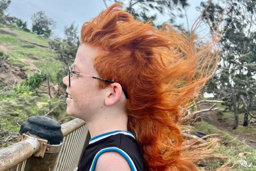 A side profile of a young boy with glasses with a long red mullet blowing in the wind