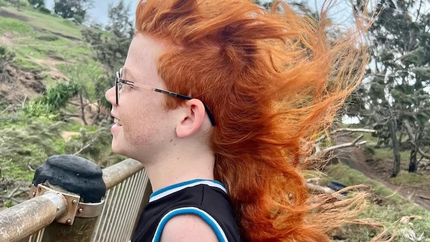 A side profile of a young boy with glasses with a long red mullet, which is blowing in the wind.