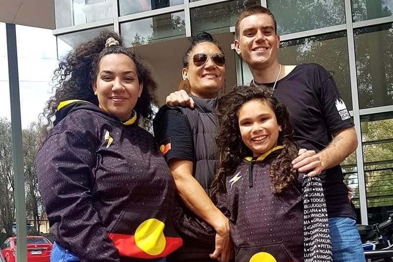 A smiling woman with three smiling children aged from early teens to late teens in clothes carrying the Aboriginal flag