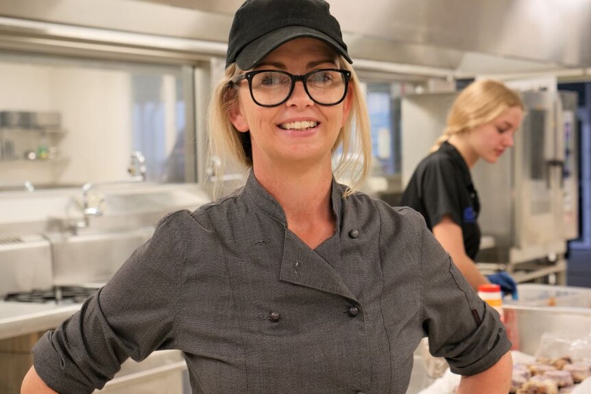 Woman in cap and glasses stands in industrial kitchen.