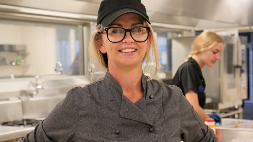 Woman in cap and glasses stands in industrial kitchen.