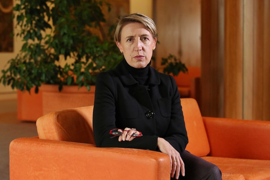 A woman wearing all black sits on an orang couch. Her arms are folded across her chest and she is holding a red pair of glasses.