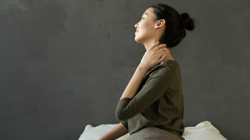 a woman sits up straight on a bed holding her neck