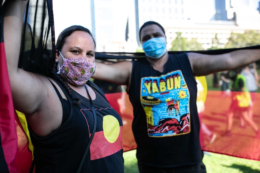 two indigenous women wearing masks and holding the Aboriginal flag behind them in a park