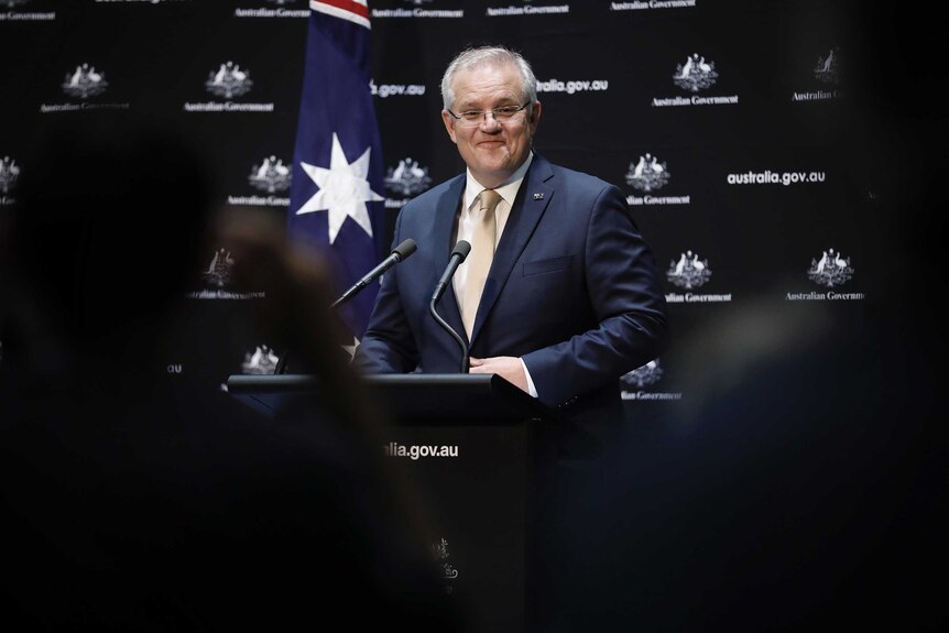 Morrison smiles at journalists. He's standing at a lectern in front of black and white Australian Government signage.