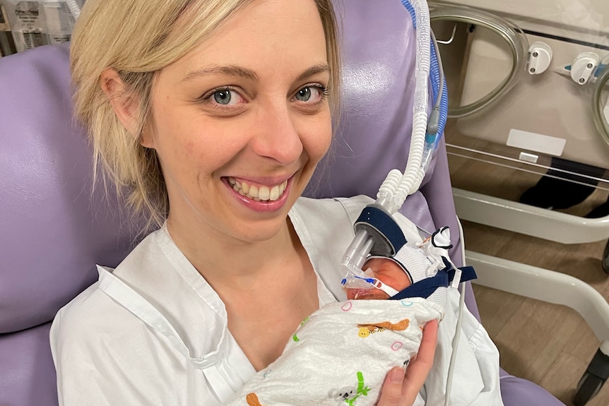 An image of Erin sitting down in hospital gowns holding her premature baby 