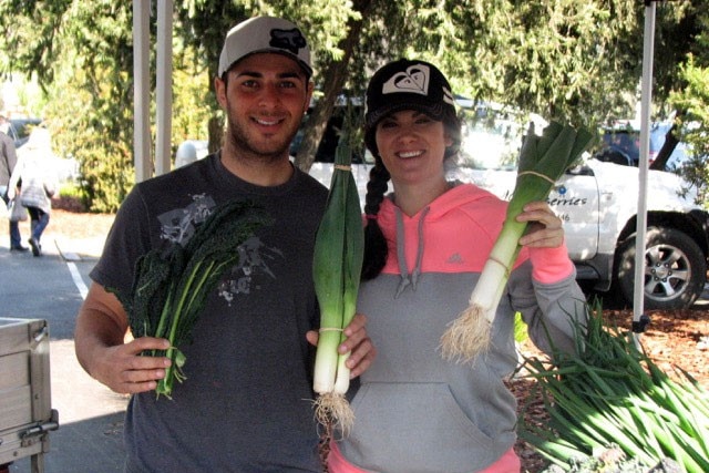A picture of a young man and woman holding leeks at a farmers' market stall.
