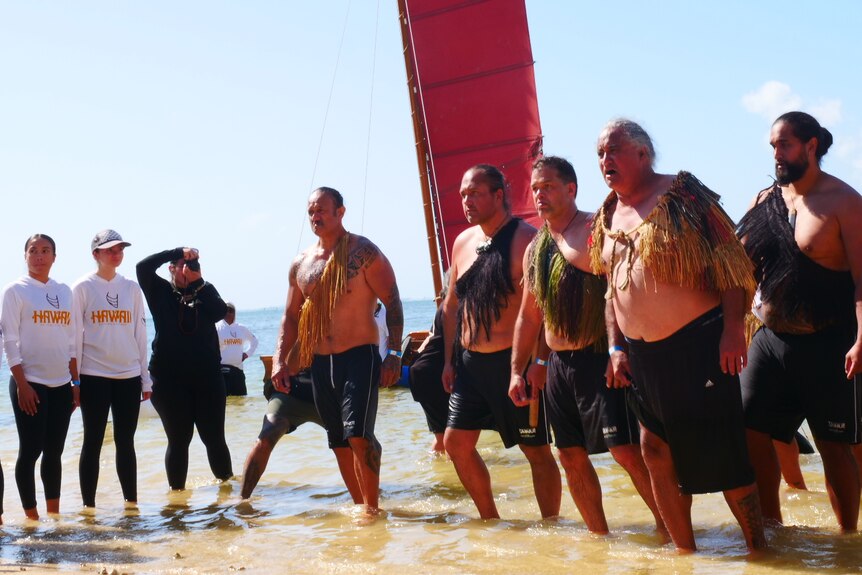 A group of men standing in water perform a haka while others watch.