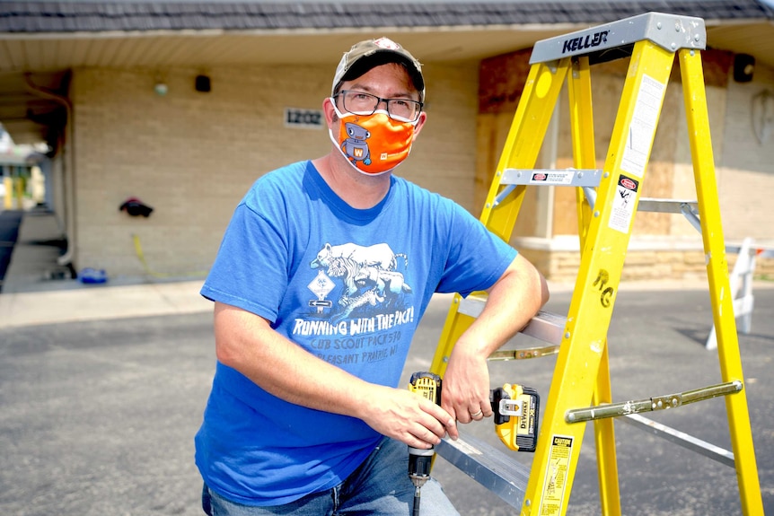 A man in a face mask leans against a ladder