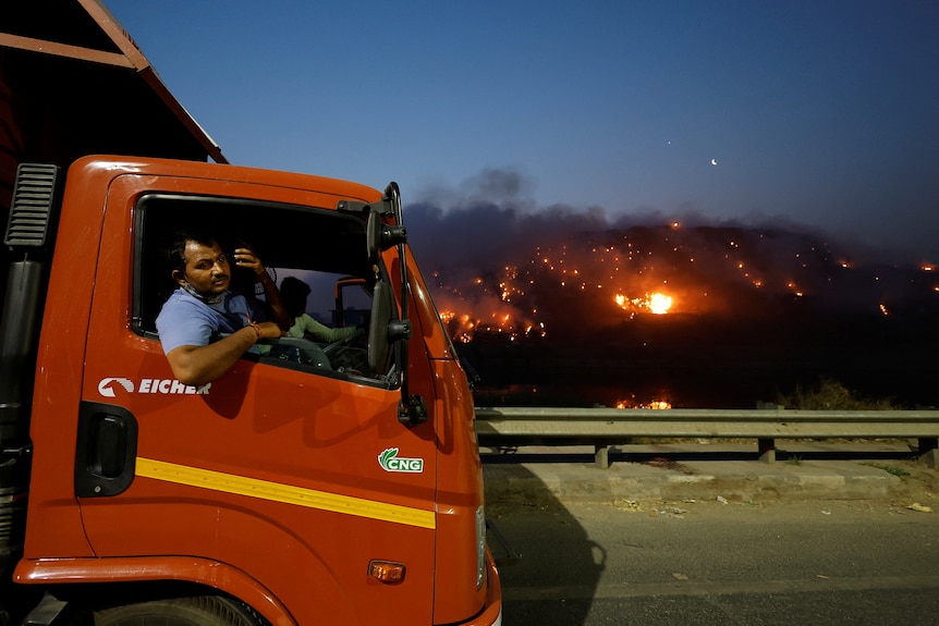 A truck driver talks on his mobile phone as smoke billows from burning garbage