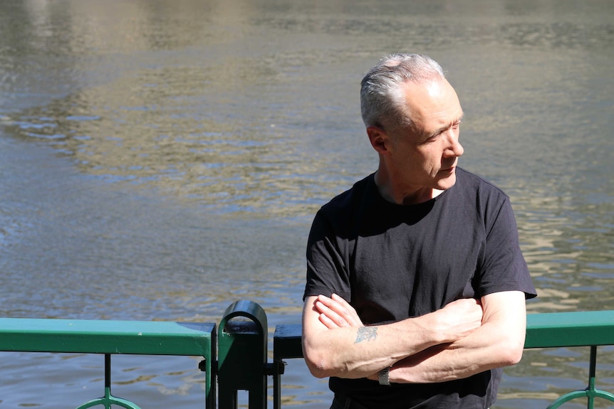 Before a backdrop of water, a man stands against a green railing in black t-shirt, with arms folded, looking sidewards.