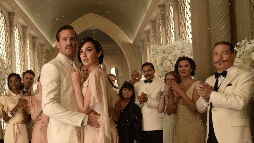 A group of people in black tie dress clap as they surround a troubled looking bride and groom in an ornate space