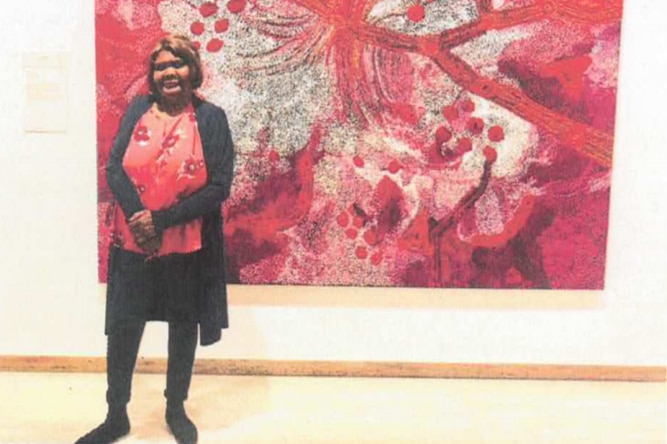 Sylvia Ken stands in front of a predominantly red painting. She wears a red shirt, black trousers and black cardigan.