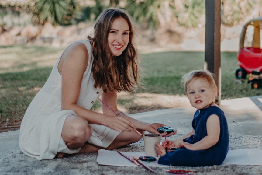 India Gladwood sitting and smiling on a picnic rug, with her small toddler son sitting next to her in a park.