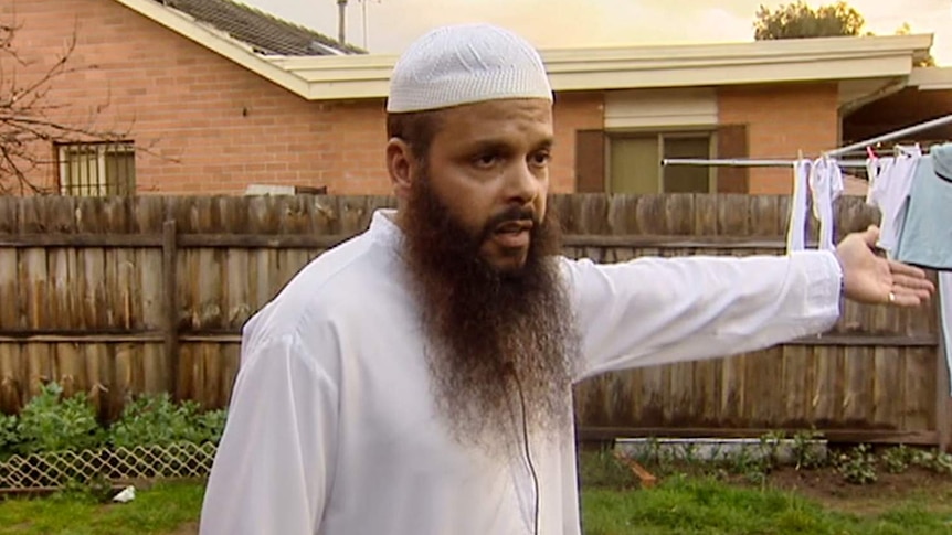 Man wearing white Islamic cap with long brown beard gestures toward washing line with fence and brick home in background