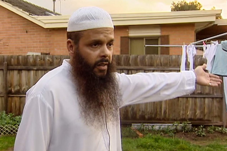 Man wearing white Islamic cap with long brown beard gestures toward washing line with fence and brick home in background