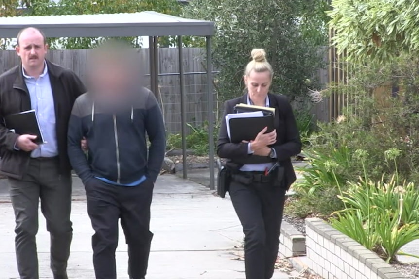 Police escort a man, whose face has been blurred, down a driveway.