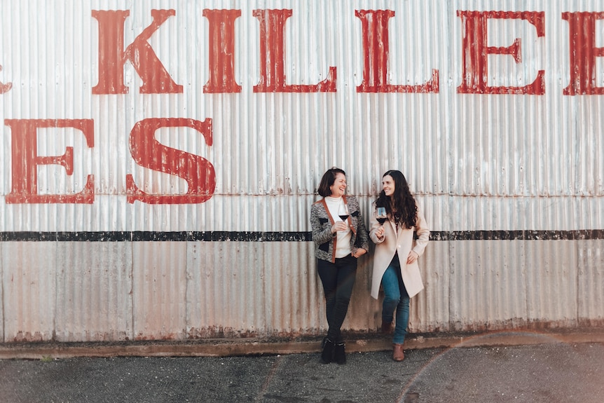 Two women lean on a shed holding wine glasses 