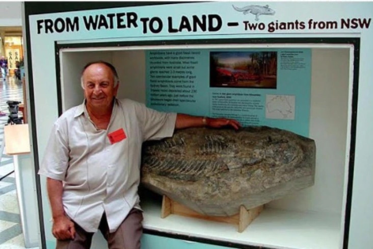 An older man wearing a white shirt sitting on a ledge wrapping his arm around a giant fossil stone