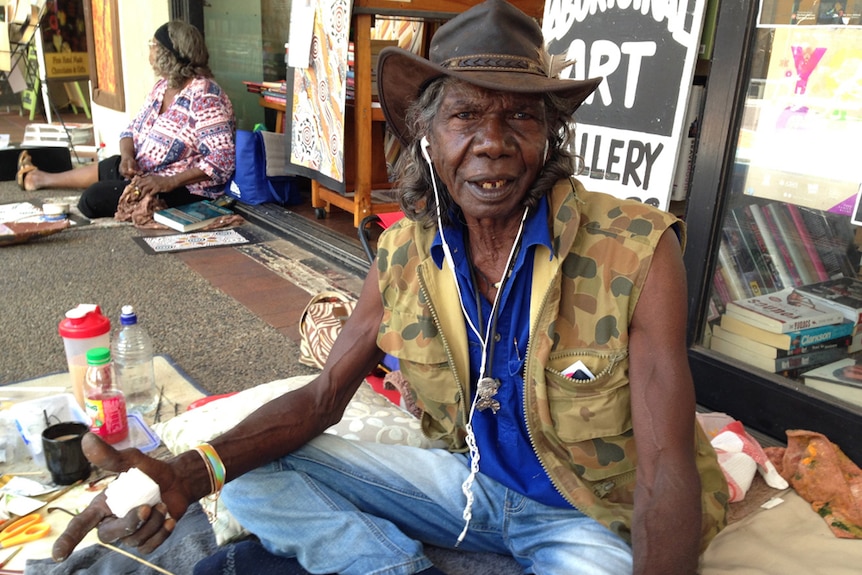A profile photo of actor David Gulpilil sitting outside a book store at the Darwin mall with canvases and paints