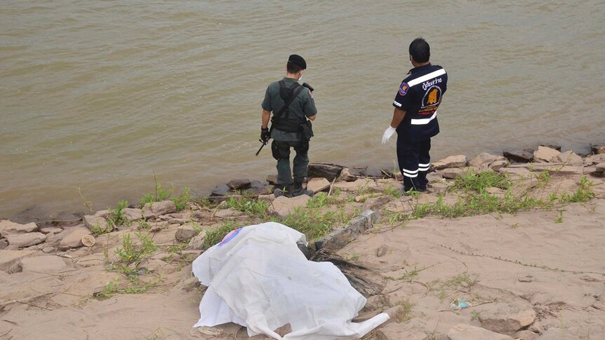 A rescuer and a police officer stand near a covered body on the shore of the Mekong River in Nakhon Phanom province, Thailand.