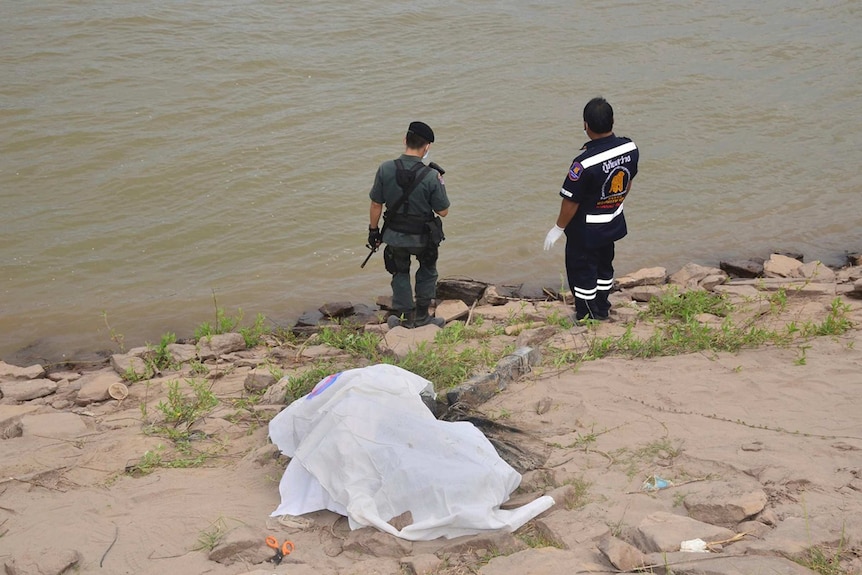 A rescuer and a police officer stand near a covered body on the shore of the Mekong River in Nakhon Phanom province, Thailand.