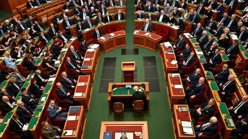 A image of the Hungarian Parliament from higher up, the room is full of people in suits sitting at desks.