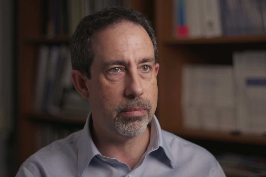 A man with a grey beard sits for a TV interview, looking past the camera with a blank expression.