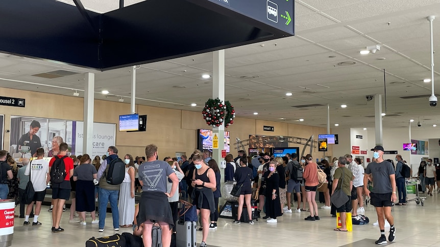 A large crowd of people milling around a baggage terminal in an airport. Most wear masks. Christmas decorations are hung up.